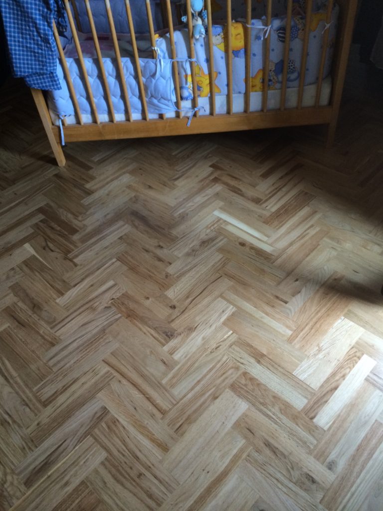 Holzboden im Fischgrätmuster, daneben ein Babybett mit buntem Bettzeug.