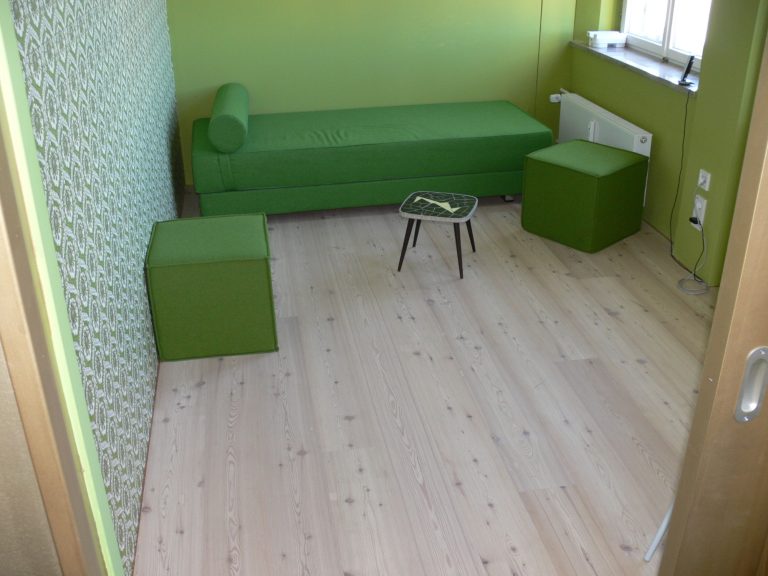 Raum mit grüner Wand, grünem Sofa, zwei grünen Hockern und hellem Holzboden.