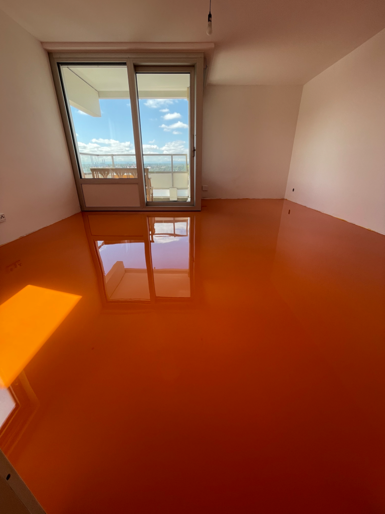 Ein Raum mit orangefarbenem Boden und großen Fenstern, die Tageslicht hereinlassen.